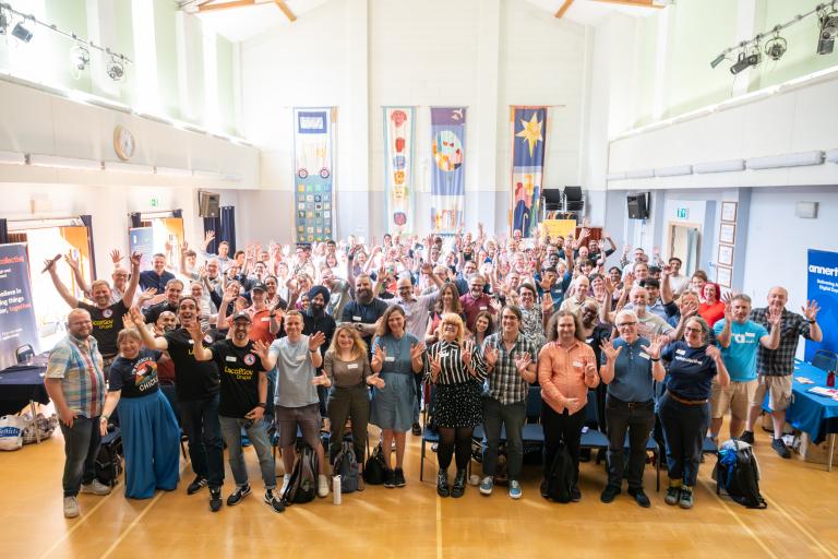 Group photo from LGD camp. Obver 100 people in a big hall, smiling with their arms raised.