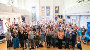 Group photo from LGD camp. Obver 100 people in a big hall, smiling with their arms raised.